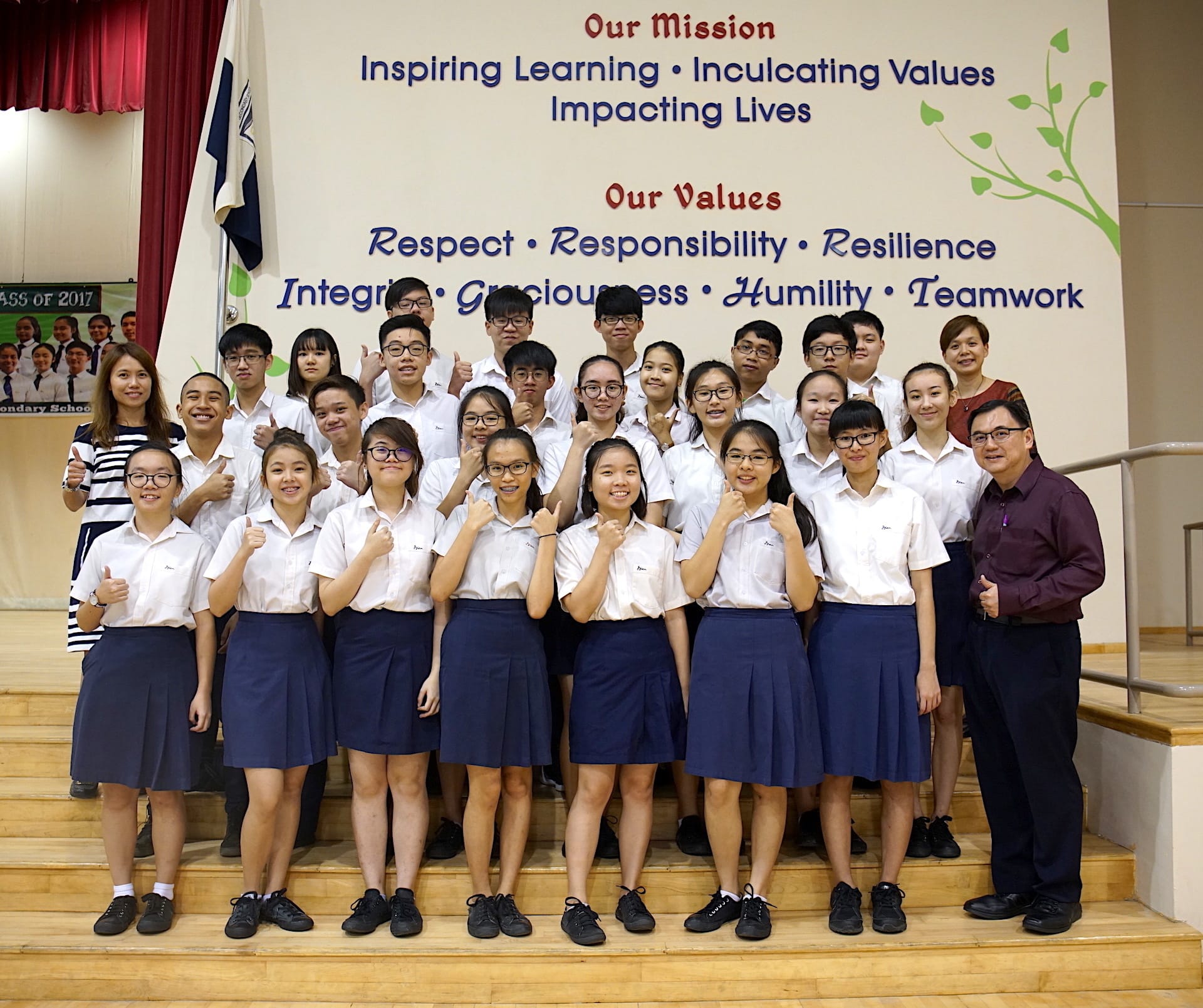 Group of school students in white and navy uniforms posing with thumbs up in front of a school mission and values banner.